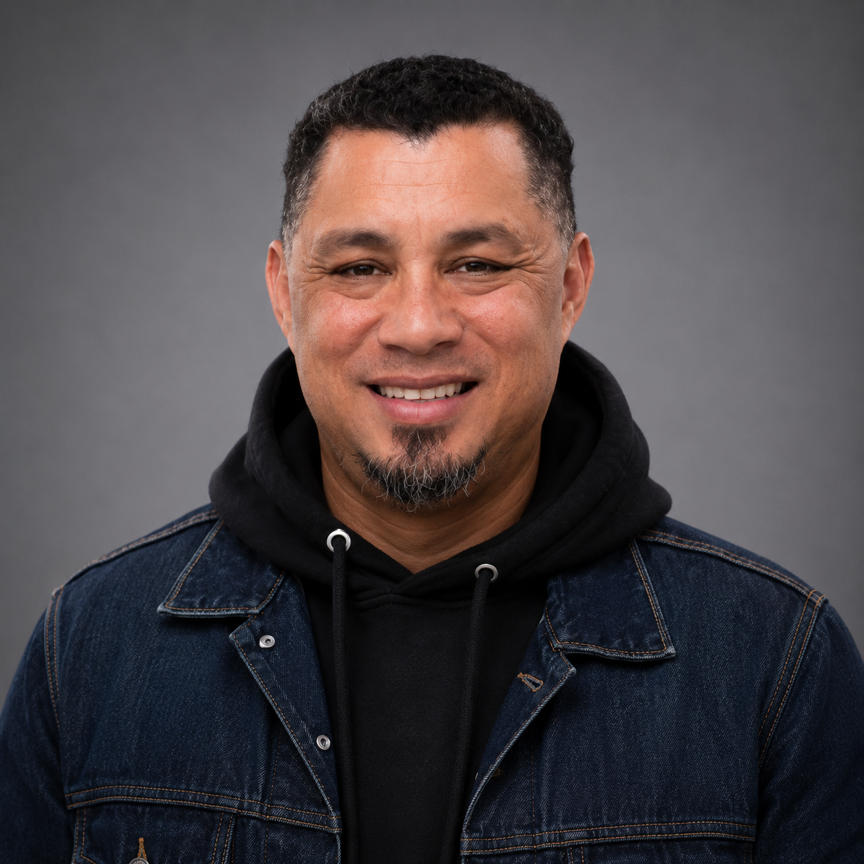 A man with short dark hair and goatee is smiling and wearing a black hoodie sweatshirt under a dark denim jacket.